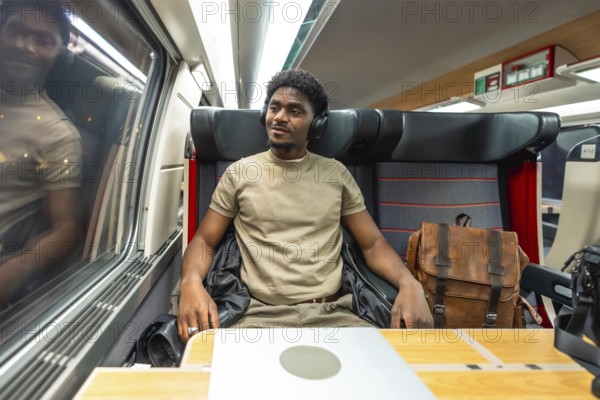 Young black man in headphones and smart casual clothes working on a laptop and gazing out the train
