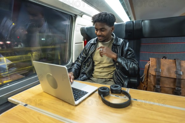 Young man using a laptop and headphones, smiling while traveling by train, reflecting modern