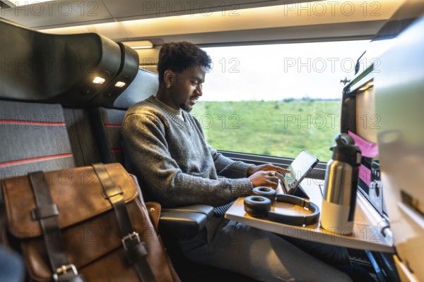 Young african businessman staying productive on a train, working on a tablet with headphones,