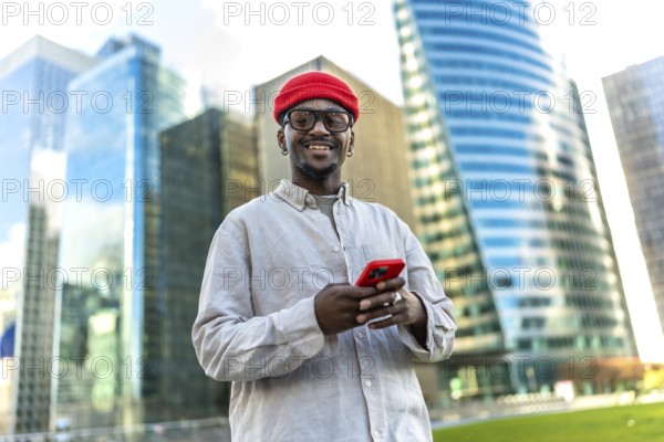 Young black man smiling, looking at the camera while using a smartphone in a modern urban district