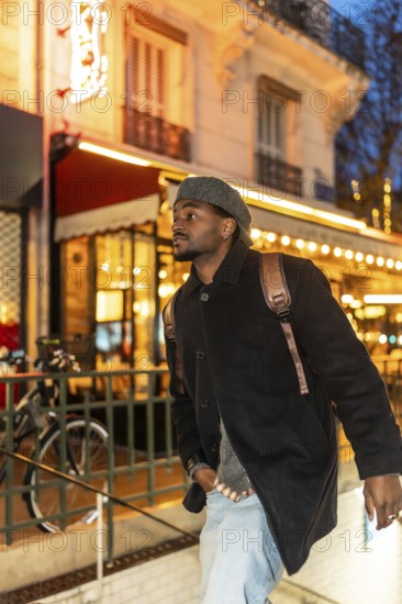 Young black man wearing a beret, coat, and backpack exploring the brightly lit streets of paris,