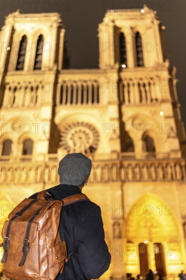 Tourist with backpack and hat seen from behind, gazing at notre dames illuminated gothic facade at
