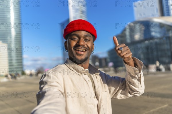 Young black man with a red beanie taking a close up selfie in an urban setting, smiling and giving
