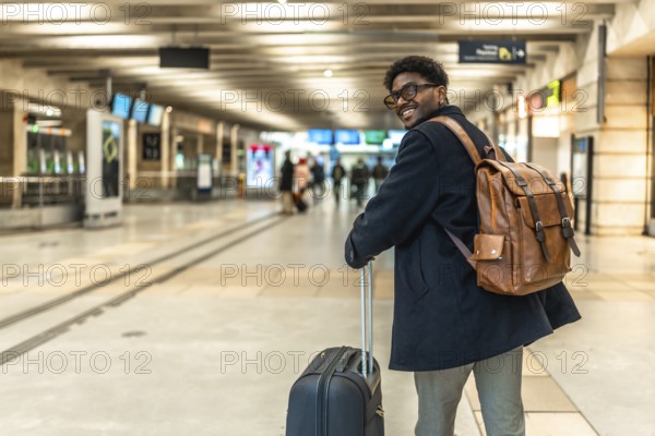 Smiling african man carrying backpack and pulling luggage, looking back while walking through a