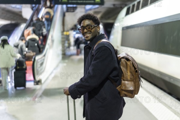 Smiling black man stands on a modern train platform with suitcase and backpack, ready for a