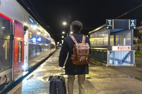 Man in coat and backpack pulls rolling luggage on a dim train platform at night, boarding for a