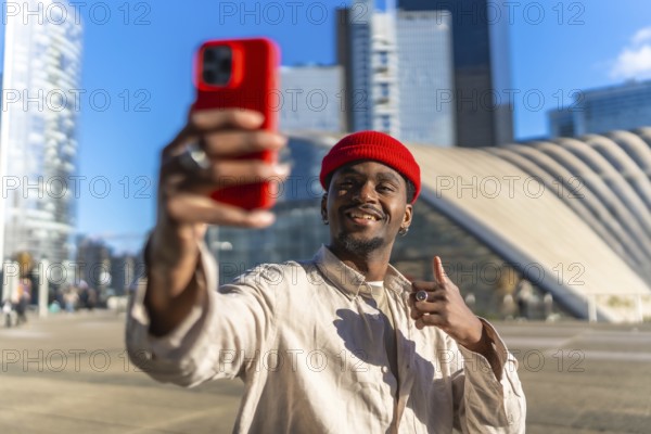 Young black man smiling while taking a selfie with a red smartphone and giving a thumbs up gesture,