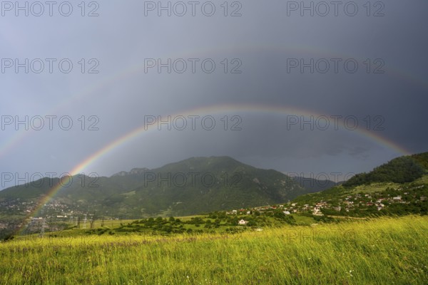 Two rainbows stretch across a green landscape with mountains and a village under a cloudy sky,