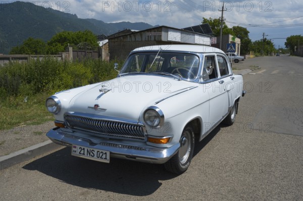 White vintage car on a country road with mountain backdrop and blue sky in summer, GAZ M-21 Volga,