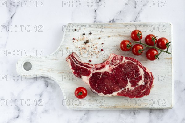 Raw rib eye steak, cowboy steak, on a white wooden chopping board, marble table, top view, no