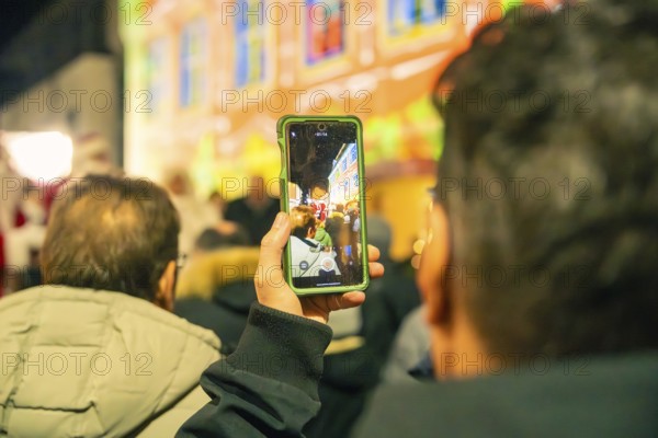 People watch a colorful event, with some capturing the scene with smartphones, Christmas market