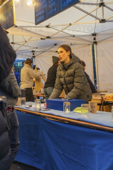 A woman in a winter coat sells goods at a market stall with a blue table, Aidlingen Christmas