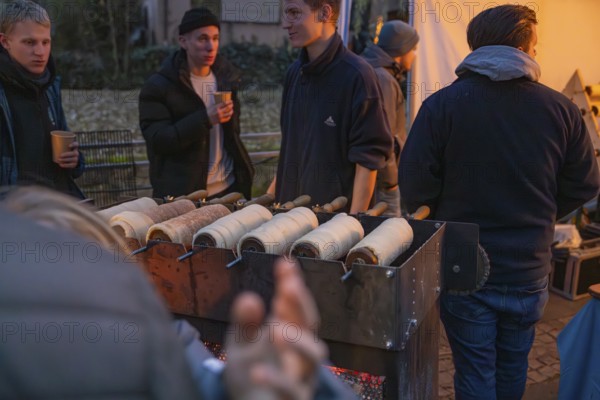 Vendors at a stand at a Christmas market prepare traditional tree striezel under evening lighting,