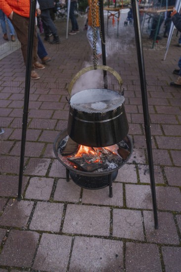 A cauldron hangs over a glowing fire on outdoor cobblestones at a Christmas market, Aidlingen