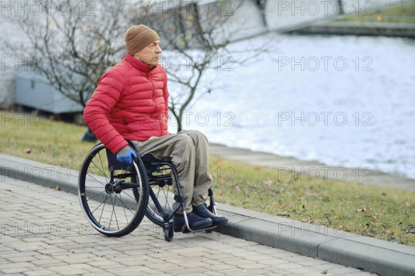 A man in a wheelchair stopped by a canal in a city park and looks at the water. He wears a red down
