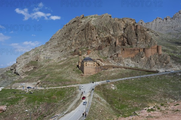 A fortress on a mountain with a road and cars in the foreground under a clear blue sky, Eski