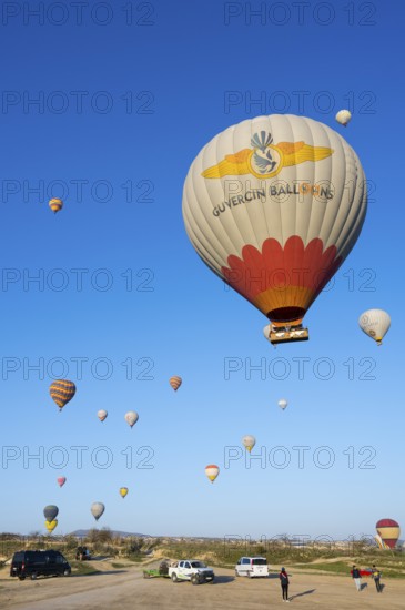 Hot air balloons rise from a meadow while people and cars are on the ground, Göreme National Park,