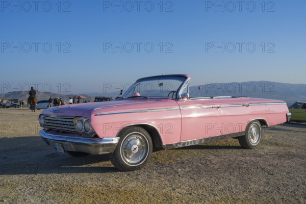 A pink vintage convertible car stands in a vast landscape with mountains in the background and blue
