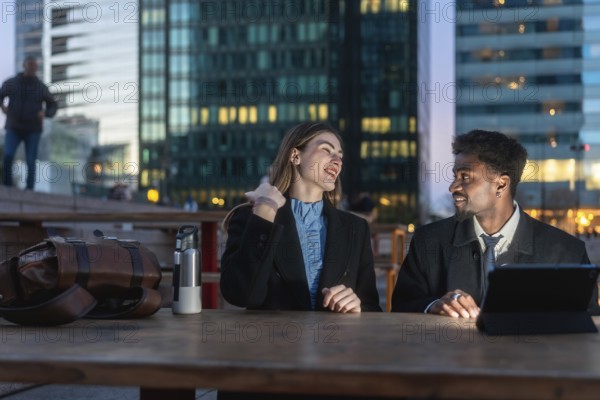 Diverse business colleagues having an outdoor conversation at dusk, sitting at a table in a modern