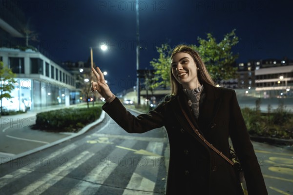 Young woman standing on an urban street at night, smiling and making a peace sign gesture while