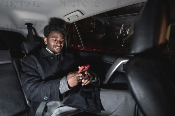Young black man relaxed in a ride share backseat at night, smiling and browsing content on his