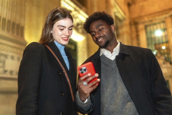 Diverse couple standing close, looking at a smartphone screen and smiling, enjoying connecting and