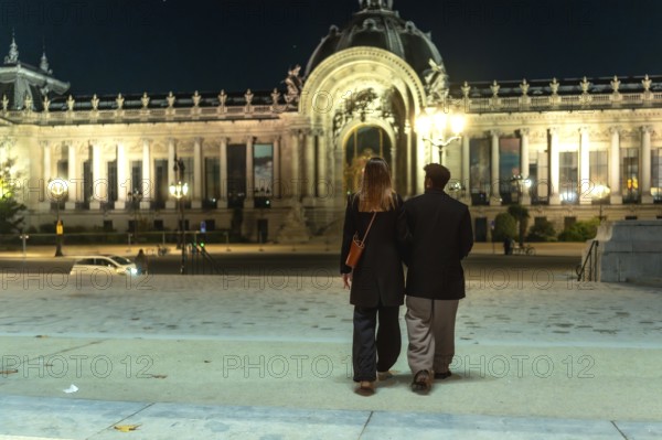 Couple walking arm in arm beneath the illuminated facades of petit palais at night, enjoying a
