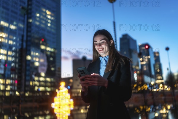 Young professional woman standing in a downtown night scene, smiling as her smartphone glows while
