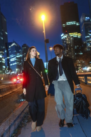 Diverse business couple walking with luggage along a downtown sidewalk at night, passing