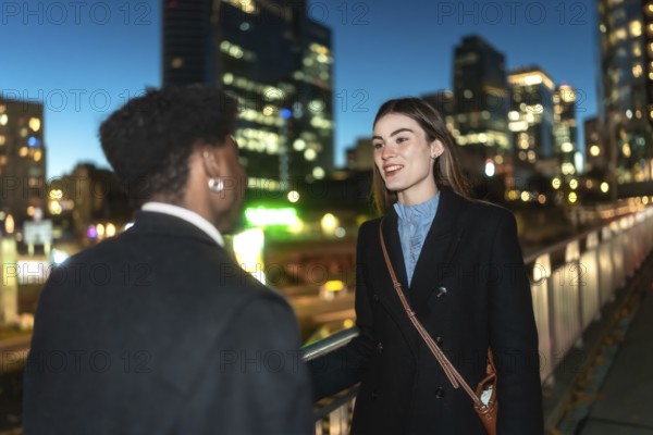 Two colleagues converse warmly on an urban pedestrian bridge at night, city lights and blurred