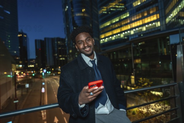 Young black businessman standing on a bridge, holding a modern smartphone with a red case, smiling