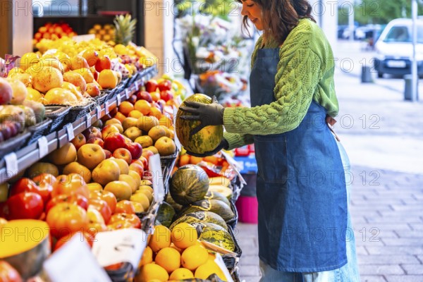 Female salesperson wearing a denim apron and gloves, arranging cantaloupe melons on a colorful
