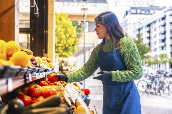 Young greengrocer woman wearing an apron and gloves arranging fresh organic fruits and vegetables