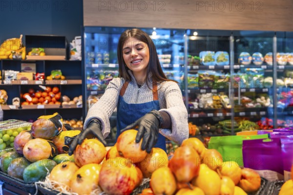 Smiling young supermarket employee in apron and gloves arranging fresh pomegranates, mangos and
