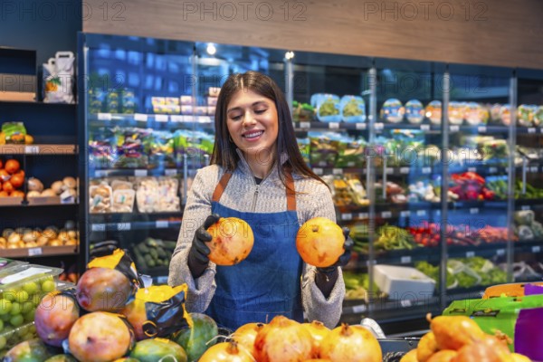 Female grocery store employee wearing an apron and gloves, smiling while holding two pomegranates,