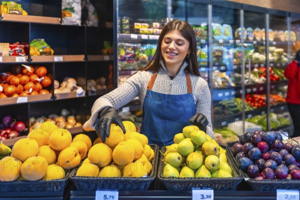 Woman grocer wearing gloves and an apron, smiling while arranging fresh produce like apricots,