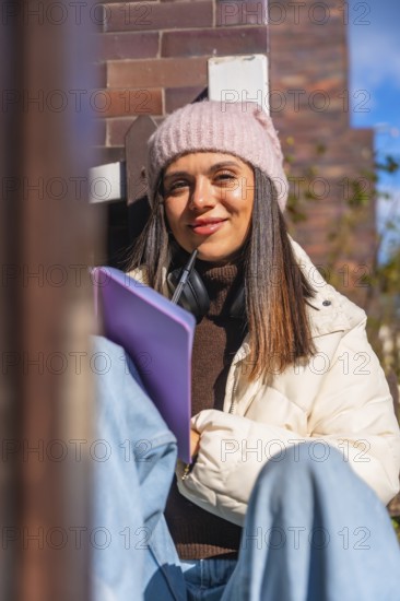 Woman student smiling and looking at the camera while writing in a journal outdoors, wearing a pink