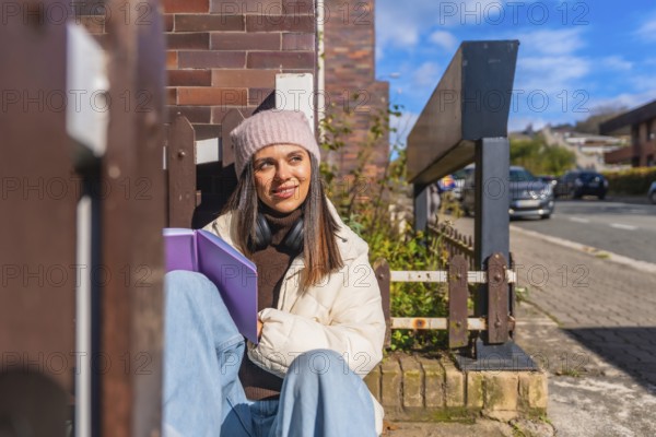 Young woman sitting outdoors on a sunny day, wearing a beanie and headphones, holding a book,