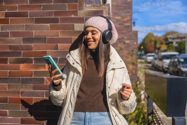 Woman smiling, wearing headphones and a beanie, holding a smartphone, standing against a brick wall