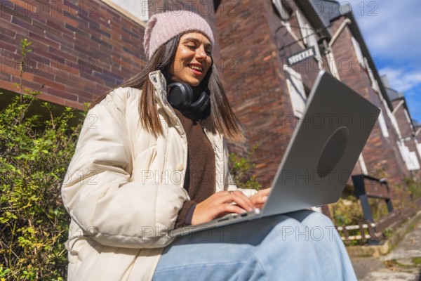 Young woman smiling and typing on a laptop, dressed in winter clothes, sitting outside a brick