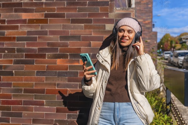 Young woman leaning against a brick wall, smiling with wireless headphones and smartphone, enjoying