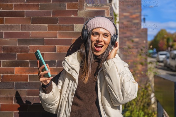 Young woman singing along to music on wireless headphones, holding her phone against a brick wall