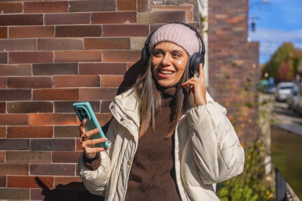 Young woman smiling while enjoying music through her headphones, holding a smartphone outdoors