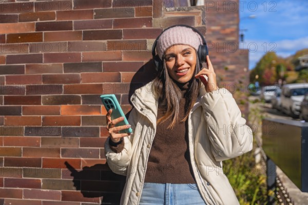 Smiling young woman dressed in warm winter clothes standing by a brick wall, peacefully listening