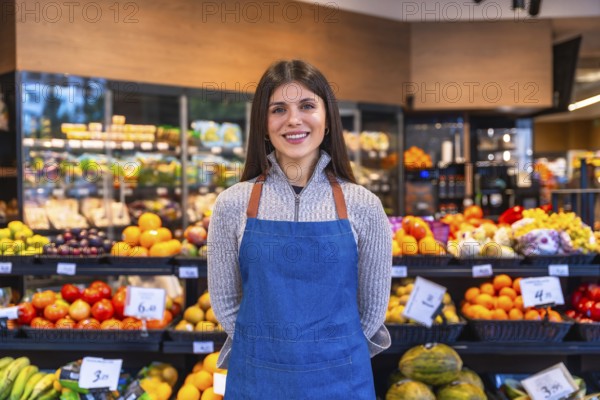 Woman wearing apron standing in a grocery store aisle, smiling at the camera, surrounded by fresh