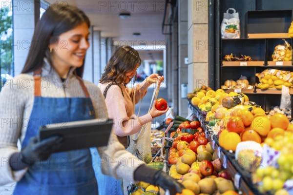Greengrocer smiling and holding a digital tablet while a happy woman consumer chooses fresh