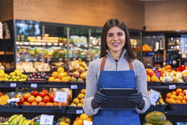 Smiling woman wearing an apron and gloves handling a digital tablet in a grocery store,