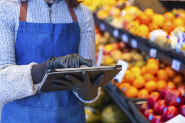 Grocery store worker wearing protective gloves and apron, using a digital tablet to manage fruit