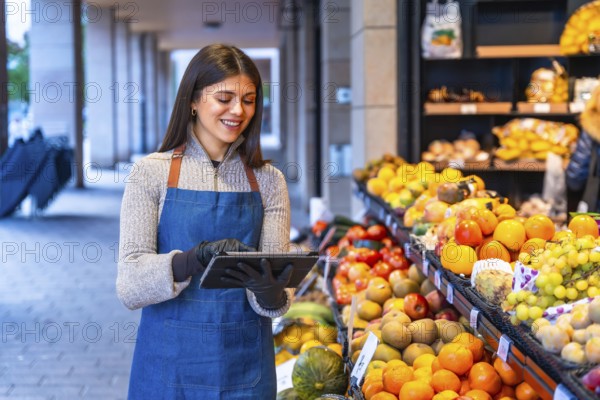 Smiling woman grocer in apron and gloves uses a tablet to manage inventory and orders at a fresh