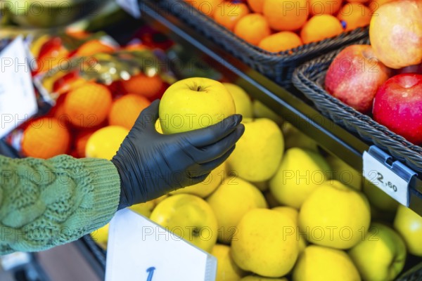 Hand in a black protective glove selects a fresh yellow apple from a supermarket produce display,